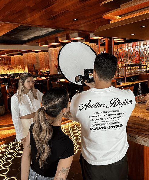 Behind-the-scenes photo shoot inside a stylish restaurant, with a model posing and a photographer adjusting lighting and camera setup